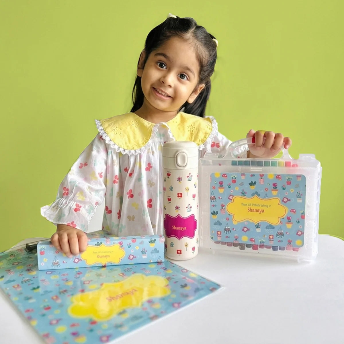 A young girl smiles while holding a personalized stationery set and a insulated water bottle. The set includes a oil pastels, a box dom pencils, and an acrylic exam clipboard in floral design