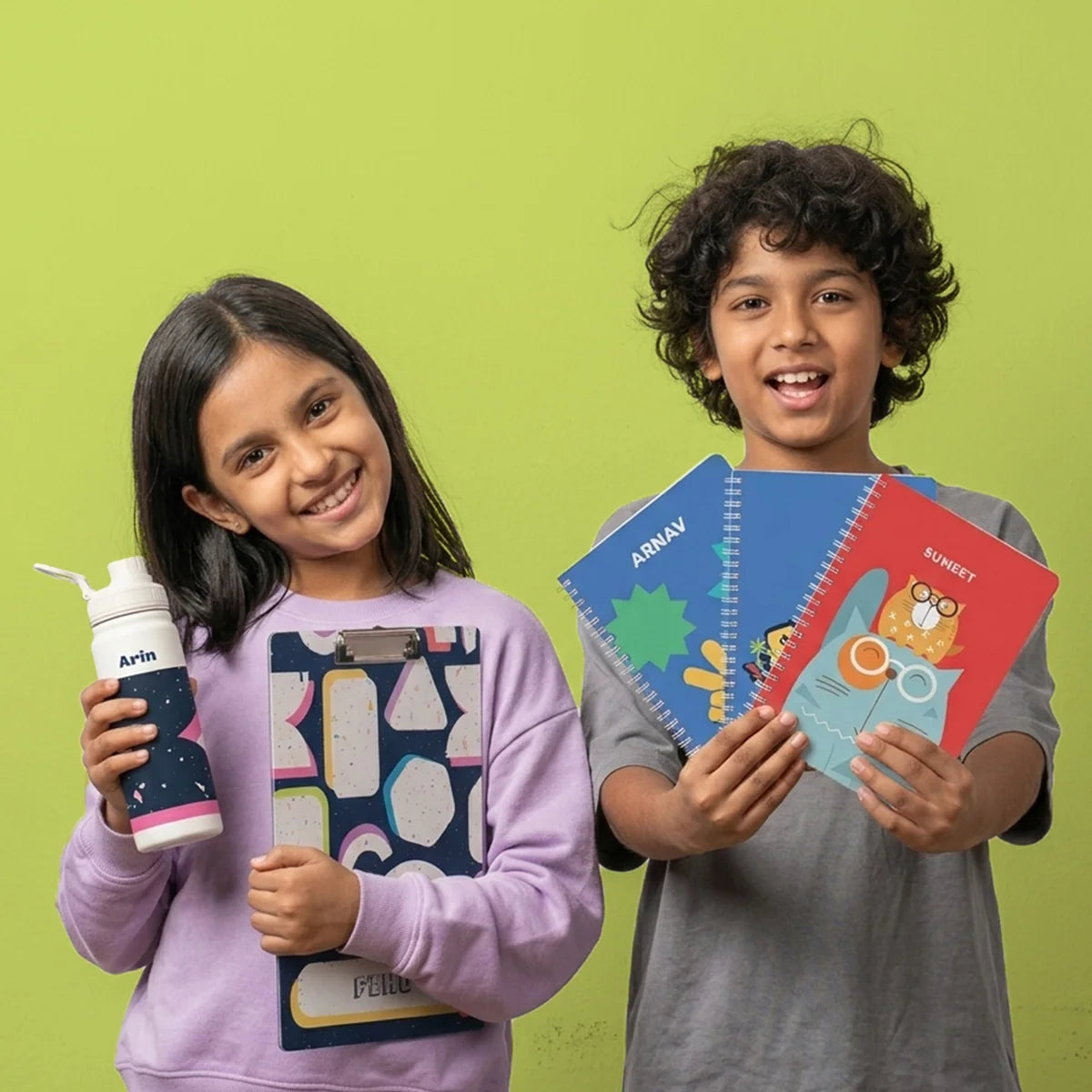Two children standing side by side holding custom return gifts. The girl on the left holds a personalized insulated bottle with a terazzo pattern and a matching exam clipboard. The boy is holding personalzied A5 size spiral notebooks.