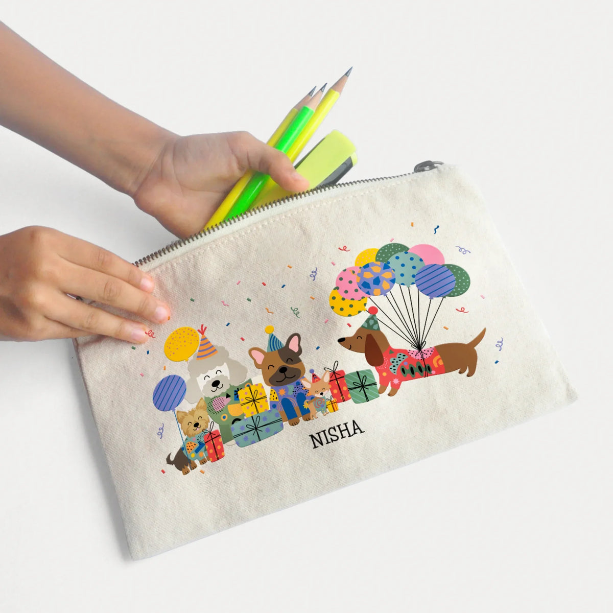 A personalized canvas pencil pouch with a colorful dog illustration and the name printed on it, held by hands holding green and yellow pencils against a clean white background.