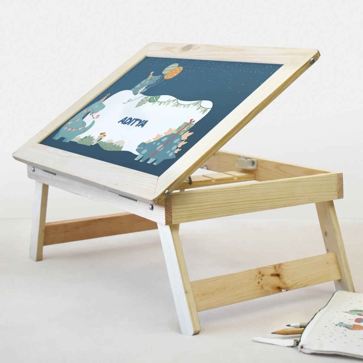 A portable, folding wooden children's drawing desk with an adjustable tabletop and a dark blue chalkboard featuring a dinosaur illustration and personalized with a name.