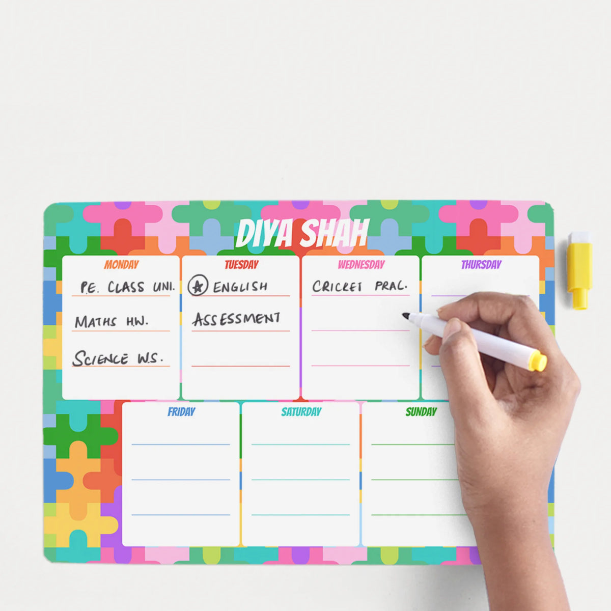Areusable,, magnetic, A4 size weekly planner whiteboard with a colorful puzzle-piece border and labeled days from Monday to Sunday. A hand is writing on the board with a white marker that has a yellow cap.