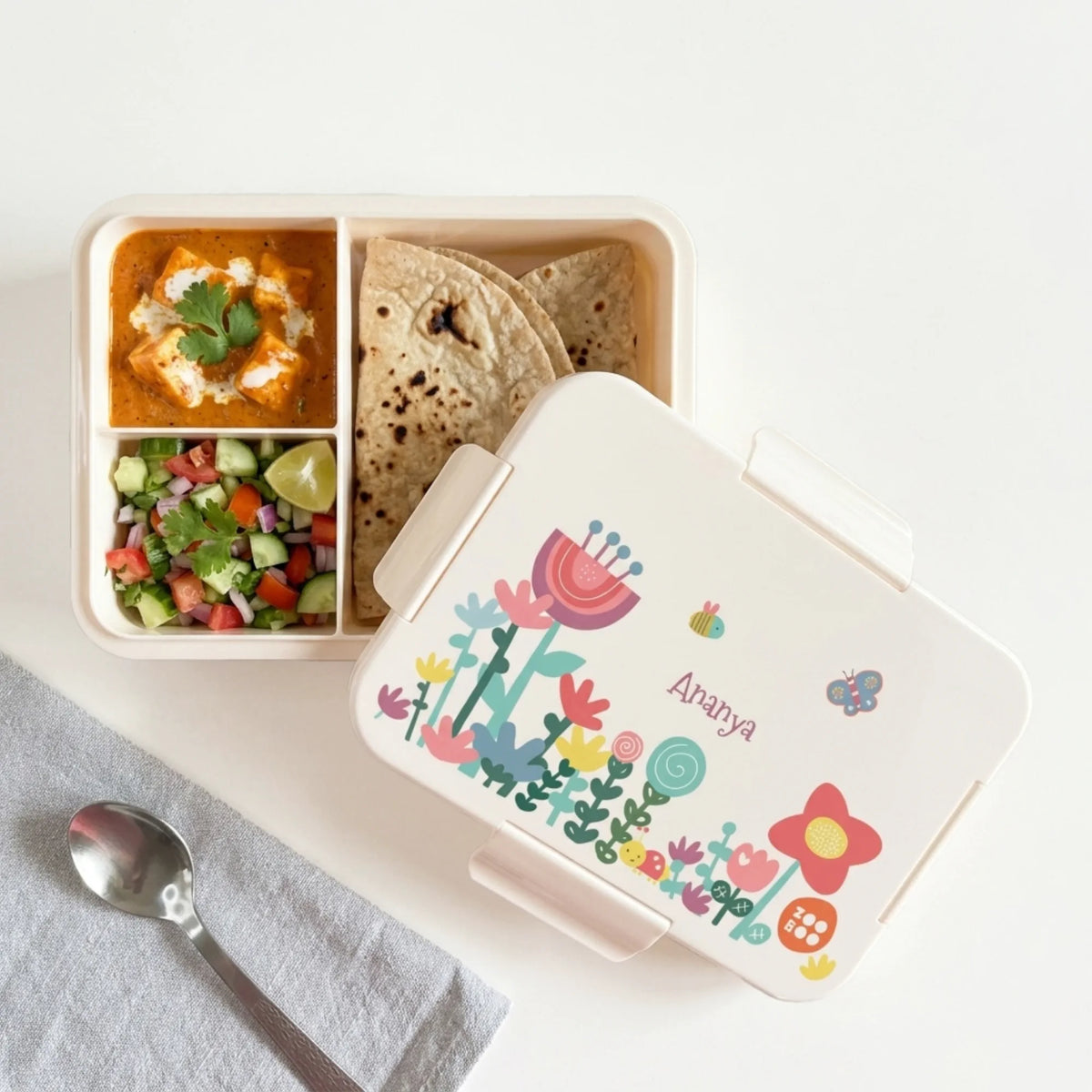 A personalized bento lunch box with a cream-colored lid decorated with colorful cartoon flowers, butterflies, and personalized with the name 'Ananya'. The box is open to reveal three-compartment, leak proof lunchbox.