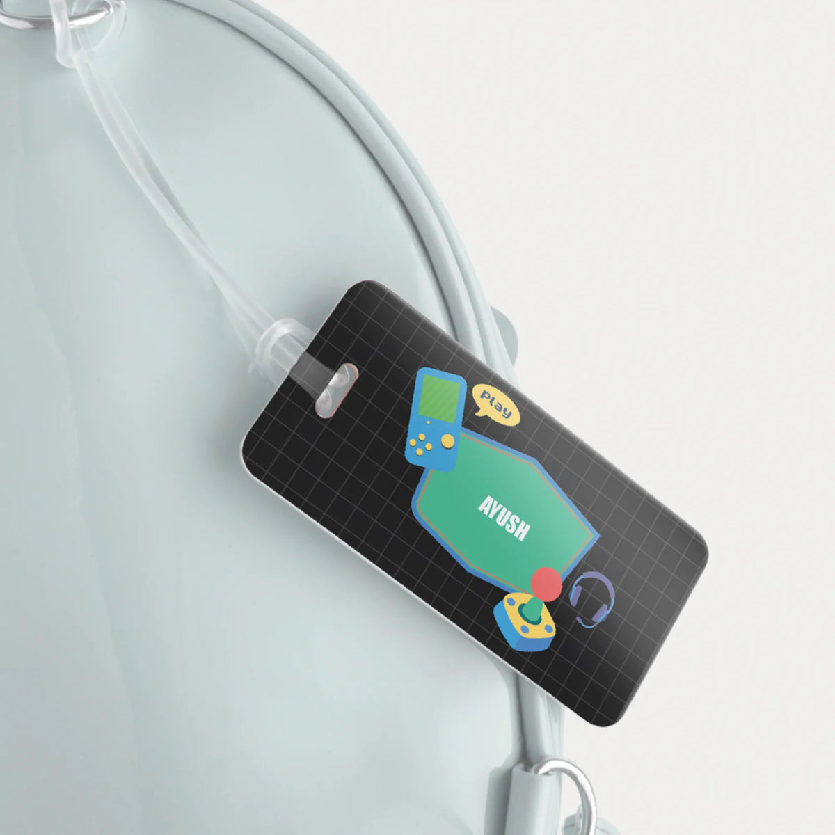A black personalized luggage tag with a grid pattern and colorful retro gaming graphics, attached to a light blue bag strap against a clean white background.