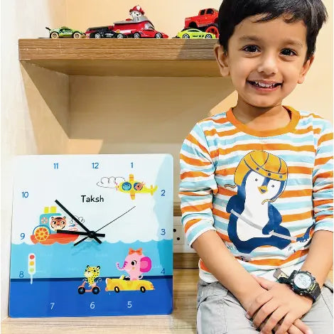 A smiling young boy sits next to a personalized children's wall clock featuring animals and vehicles.